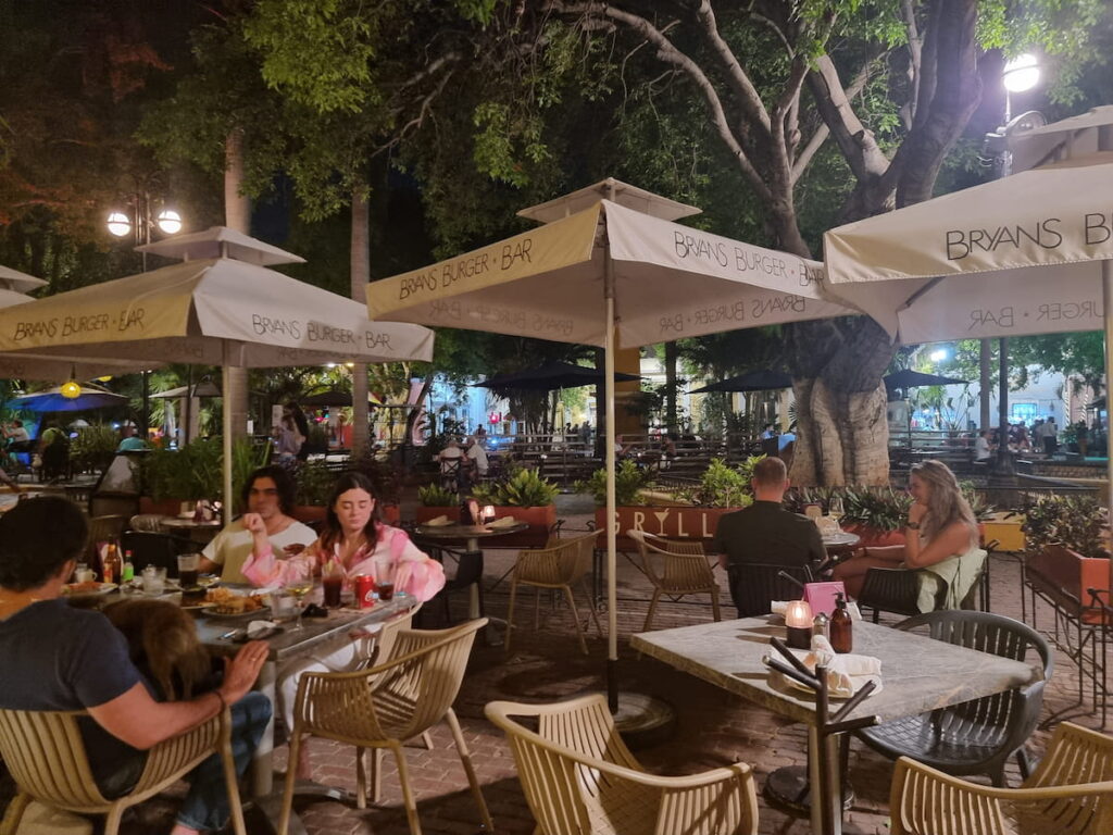 People eating outside under trees and shade umbrellas on the plaza of Parque Santa Lucia in Mérida, Mexico