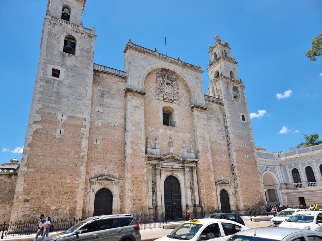 Front stone facade of Catedral de San Ildefonso in Mérida, Mexico with cars and taxis passing in front.