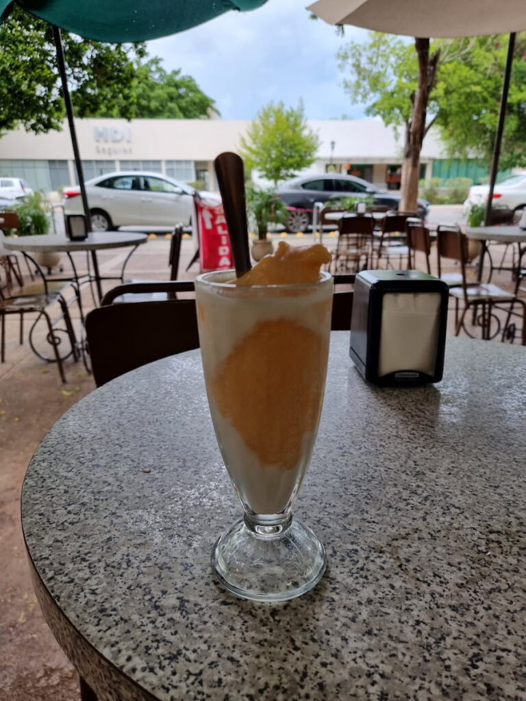 A mango champola in a tall glass dish with a spoon sits on the table at an outdoor cafe with cars and a street in the background.