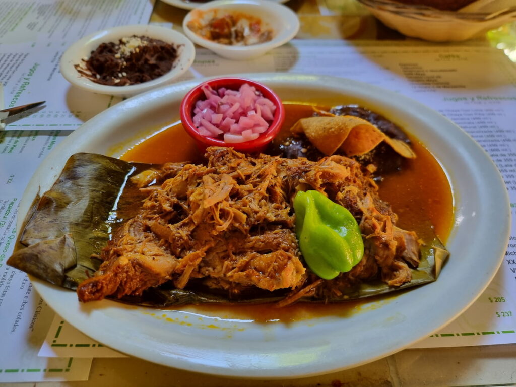 Plate of cooked cochinita pibil on top of the banana leaf with a pepper, preserved chopped onions, refried beans and some side dishes in the background.