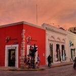 Intersection of Calle 55 and Calle 60 in Mérida, Mexico. A horsedrawn carriage waits on one street and people wait to cross as the traffic passes by. A coral colored colonial building centers the frame with other shops lining the street. The sky is lit with a rose tone from the setting sun.