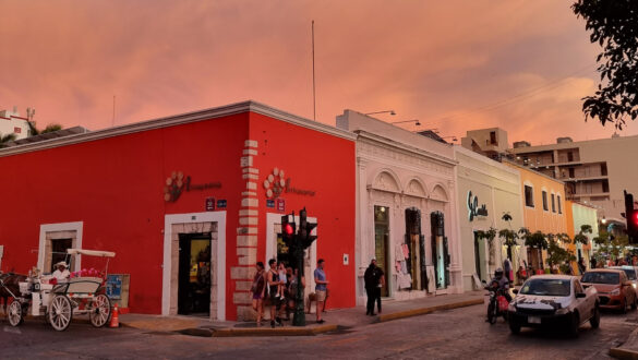 Intersection of Calle 55 and Calle 60 in Mérida, Mexico. A horsedrawn carriage waits on one street and people wait to cross as the traffic passes by. A coral colored colonial building centers the frame with other shops lining the street. The sky is lit with a rose tone from the setting sun.