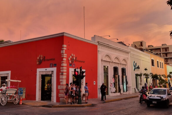 Intersection of Calle 55 and Calle 60 in Mérida, Mexico. A horsedrawn carriage waits on one street and people wait to cross as the traffic passes by. A coral colored colonial building centers the frame with other shops lining the street. The sky is lit with a rose tone from the setting sun.