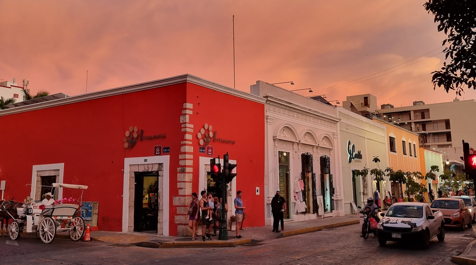 Intersection of Calle 55 and Calle 60 in Mérida, Mexico. A horsedrawn carriage waits on one street and people wait to cross as the traffic passes by. A coral colored colonial building centers the frame with other shops lining the street. The sky is lit with a rose tone from the setting sun.